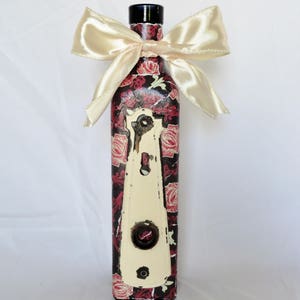 May include: A decorative glass bottle with a black top and a floral design featuring red roses and a white keyhole. The bottle is tied with a cream-colored satin ribbon.