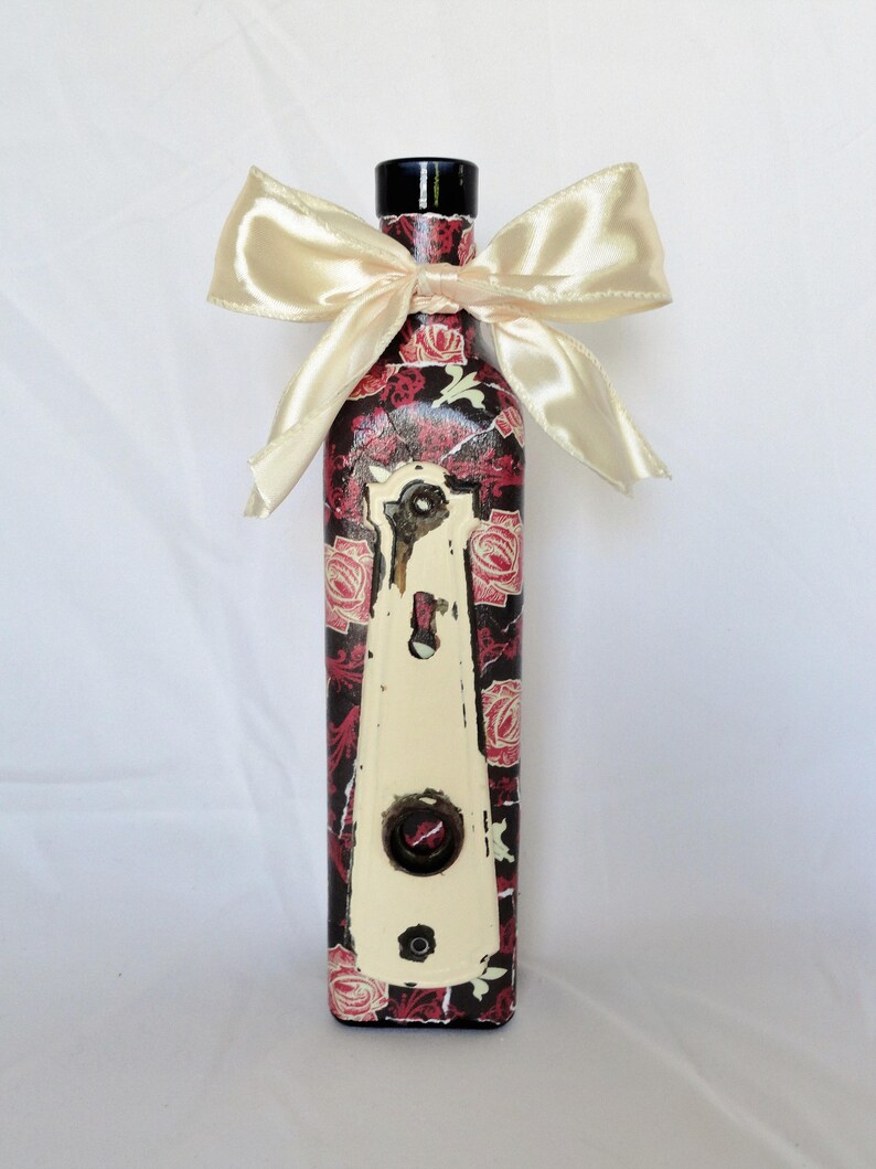 May include: A decorative glass bottle with a black top and a floral design featuring red roses and a white keyhole. The bottle is tied with a cream-colored satin ribbon.