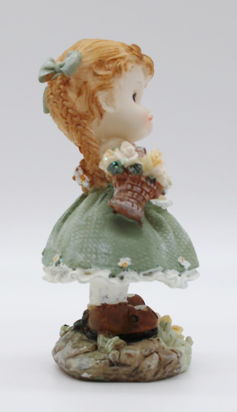 Op de afbeelding: Een beeldje van een meisje met bruin haar dat een groene jurk draagt en een mandje met bloemen vasthoudt. Ze staat op een stenen basis.