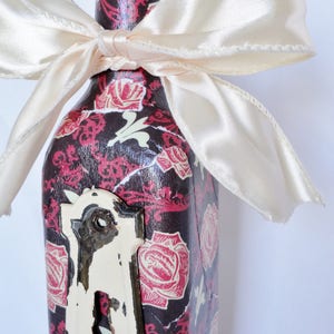 May include: A glass bottle with a black and white floral design and a white satin bow. The bottle has a vintage keyhole design on the front.