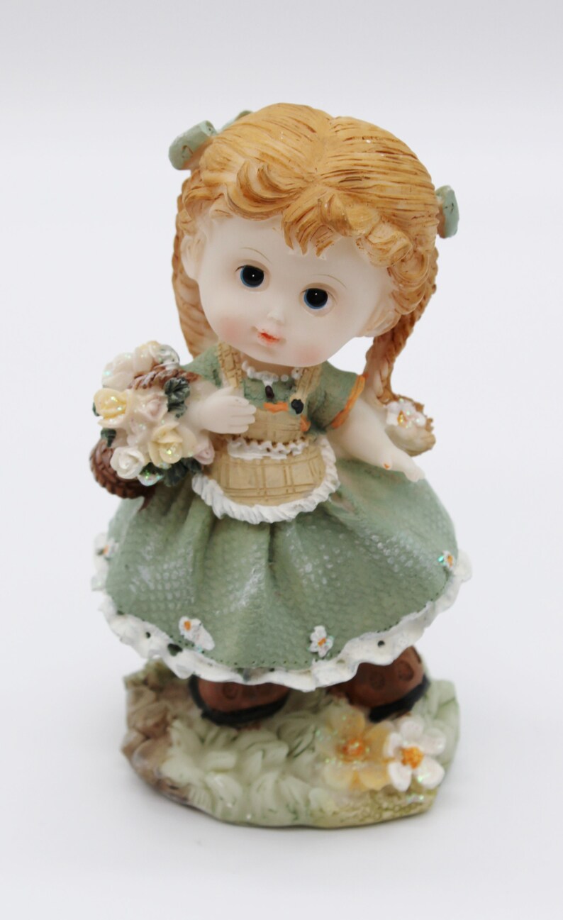 Op de afbeelding: Een beeldje van een meisje met blonde vlechten, dat een groene jurk draagt en een mandje met bloemen vasthoudt. Het beeldje staat op een voetstuk met groene bladeren en bloemen.