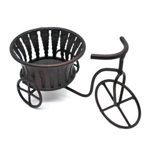 Op de afbeelding: Een decoratieve metalen plantenbak in de vorm van een driewieler met een mandje aan de voorkant. De plantenbak is donkerbruin geverfd en heeft een vintage uitstraling.