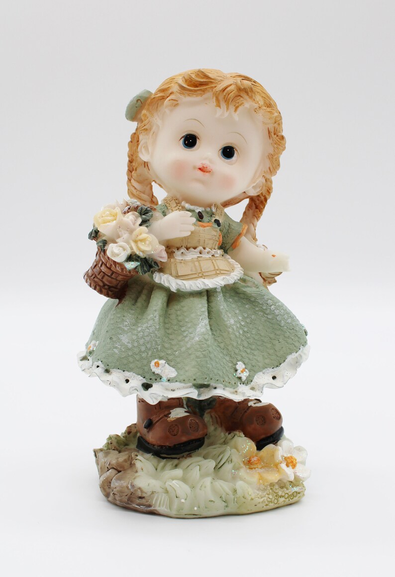 Op de afbeelding: Een beeldje van een meisje met bruin haar dat een groene jurk en bruine laarzen draagt. Ze houdt een mand met bloemen vast en staat op een voetstuk met groen gras en bloemen.