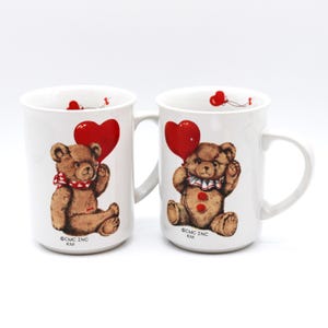Op de afbeelding: Twee witte keramische mokken met een bruine teddybeer die een rode hartballon vasthoudt. De teddyberen hebben rode en witte accenten. De mokken hebben een oor en de tekst "©CMC INC KM".