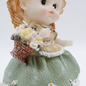 Op de afbeelding: Een porseleinen beeldje van een meisje met blonde vlechten, dat een groene jurk draagt en een mand met bloemen vasthoudt.
