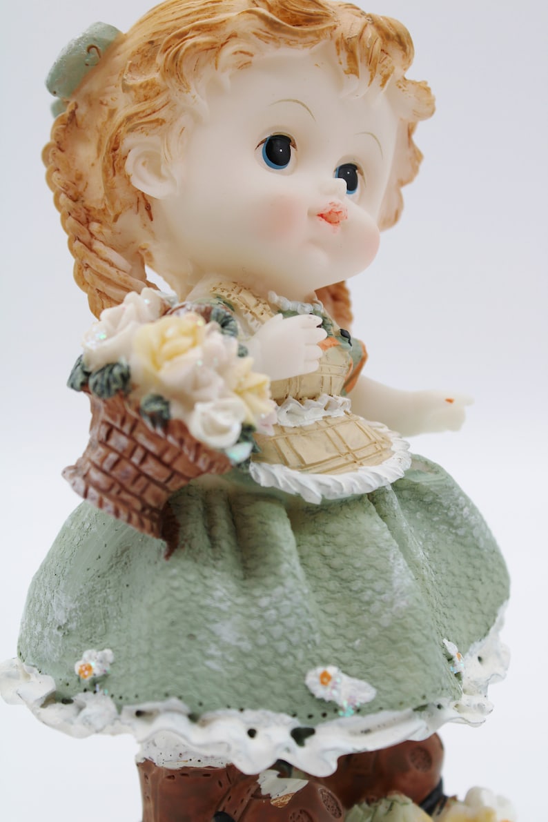 Op de afbeelding: Een porseleinen beeldje van een meisje met blonde vlechten, dat een groene jurk draagt en een mand met bloemen vasthoudt.