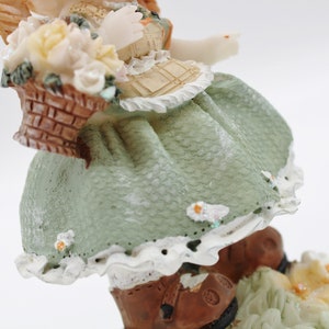Op de afbeelding: Een porseleinen beeldje van een meisje dat een groene jurk draagt met witte kant en bruine laarzen. Ze houdt een mand met witte bloemen vast en heeft engelenvleugels.