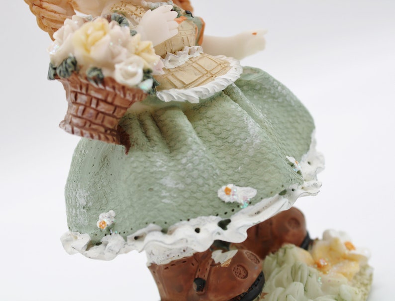 Op de afbeelding: Een porseleinen beeldje van een meisje dat een groene jurk draagt met witte kant en bruine laarzen. Ze houdt een mand met witte bloemen vast en heeft engelenvleugels.
