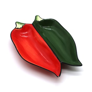 Op de afbeelding: Een keramische serveerschaal in de vorm van twee aan elkaar verbonden chilipepers. Eén peper is felrood, de andere groen. Beide hebben zwarte contouren en een glanzende afwerking. Ontworpen voor het serveren van snacks of dips.