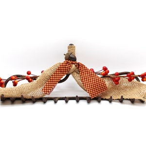 May include: A rustic metal rake decorated with burlap and a red and white checkered ribbon. The rake is adorned with a sprig of red berries.