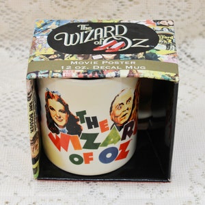 Op de afbeelding: Een witte keramische mok met een sticker van de filmposter voor "The Wizard of Oz". De mok toont Dorothy en de Tovenaar, en de tekst "The Wizard of Oz" in kleurrijke letters. De mok zit in een doos met een collage van filmposterafbeeldingen.