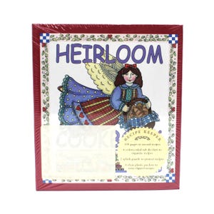 Op de afbeelding: Een rood omrand receptenboek met het woord "HEIRLOOM" bovenaan. De omslag toont een illustratie van een vrouw met vleugels die een mand vasthoudt. Tekst op de omslag geeft aan dat het 100 pagina's, verdelers, spatbeschermers en vakken heeft.