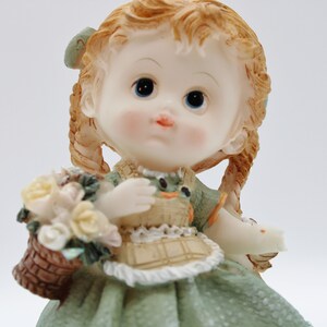 Op de afbeelding: Een porseleinen beeldje van een meisje met blonde vlechten, dat een groene jurk draagt en een mand met bloemen vasthoudt.