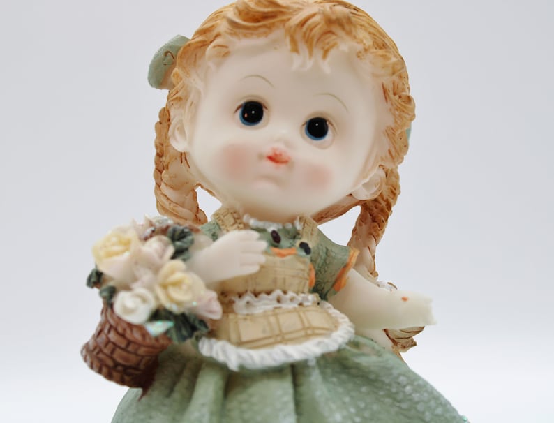 Op de afbeelding: Een porseleinen beeldje van een meisje met blonde vlechten, dat een groene jurk draagt en een mand met bloemen vasthoudt.