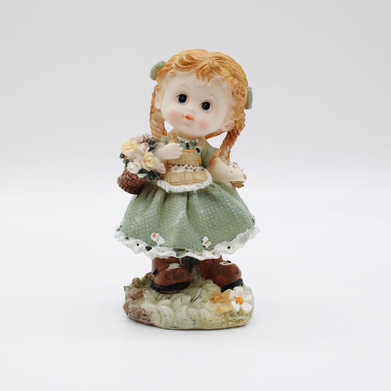 Op de afbeelding: Een porseleinen beeldje van een meisje met blonde vlechten, dat een groene jurk en bruine schoenen draagt. Ze houdt een mand met bloemen vast.