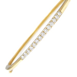 May include: A gold bangle bracelet with a row of small, clear gemstones set along the top. The bracelet is a simple, elegant design, perfect for everyday wear or special occasions. The gemstones sparkle against the gold.