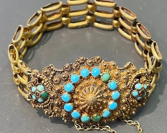 Puede incluir: Un brazalete dorado con un diseño de filigrana. La parte central presenta una disposición circular de piedras de color turquesa. El brazalete tiene una cadena de seguridad y está sobre un fondo gris.