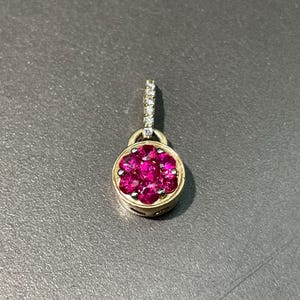 May include: A gold pendant with a cluster of vibrant pink gemstones in a circular setting. The pendant has a small loop adorned with small, clear gemstones for attaching to a chain. The pendant is set against a gray background.