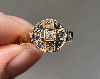 May include: A gold ring with a circular design. The ring features a central square diamond, surrounded by smaller blue gemstones and additional diamonds. The band is a simple gold color.