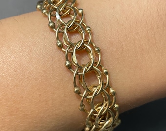 May include: A gold-colored bracelet with a woven design of interlocking loops and small, round beads. The bracelet is made of a shiny, metallic material and is displayed on a person's arm.