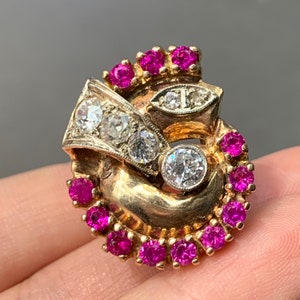 May include: A vintage gold-tone brooch with a swirling design. The brooch features three large, clear diamonds and several smaller diamonds. It is also adorned with numerous small, vibrant red rubies, creating a striking contrast.