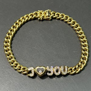 Puede incluir: Un brazalete de cadena de oro con un colgante de "Te amo" engastado con diamantes. El colgante tiene forma de corazón con un pequeño diamante en el centro.