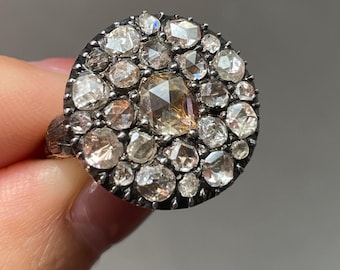May include: A silver-toned ring featuring a cluster of clear and champagne-colored gemstones. The ring's design is circular, with a central, larger stone surrounded by smaller, faceted gems. The ring is held by a hand against a neutral background.