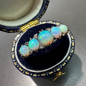 May include: A vintage brooch featuring five oval opals with iridescent colors of blue, green, and orange. The opals are set in a silver-toned metal with small accent stones. The brooch is displayed inside a blue velvet jewelry box with gold accents.