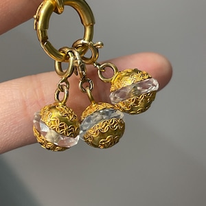 Peut inclure: Gros plan sur un collier doré avec trois breloques décoratives. Chaque breloque présente une sphère en filigrane dorée avec un centre en cristal clair et facetté. Le collier est tenu par un doigt sur un fond neutre.