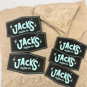 May include: Five rectangular black stickers with angled top and bottom edges. Each sticker features the light blue text Jacks Coffee Co. in a stylised font, flanked by two small four-pointed starburst icons on the left and right sides.