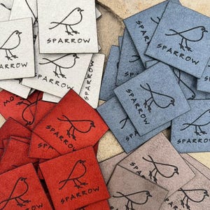 May include: A collection of square labels in red, blue, gray, and off-white colors. Each label features a black line drawing of a sparrow bird and the word SPARROW printed in black capital letters. The labels have a textured, matte finish.
