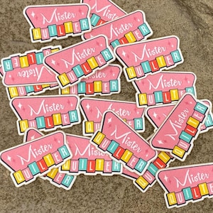 May include: A collection of die-cut stickers featuring a pink trapezoidal sign with the text Mister Quilter in white script. Below the text, the word QUILTER is displayed in individual colourful blocks of yellow, orange, red, and teal.