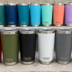Peut inclure: Une collection de treize gobelets en acier inoxydable YETI disposés sur deux rangées. Chaque gobelet possède un couvercle transparent, un bord argenté et le logo YETI gravé à la base. Les couleurs incluent vert foncé, bleu marine, bleu sarcelle, bleu vif, menthe, corail, violet, rouge, olive, noir, blanc, gris et magenta.