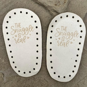 Puede incluir: Dos bases ovaladas de madera en color blanco roto para proyectos de ganchillo, cada una con una serie de agujeros preperforados a lo largo del per&iacute;metro y el texto grabado a l&aacute;ser "THE SNUGGLE IS REAL" junto a peque&ntilde;os iconos de coraz&oacute;n.