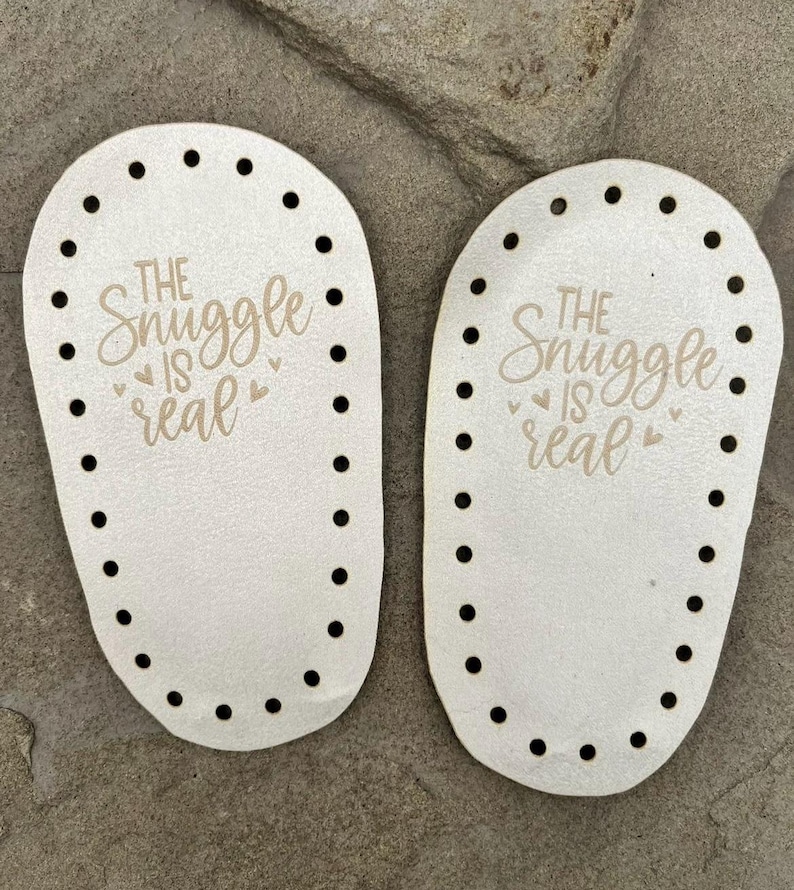 Puede incluir: Dos bases ovaladas de madera en color blanco roto para proyectos de ganchillo, cada una con una serie de agujeros preperforados a lo largo del per&iacute;metro y el texto grabado a l&aacute;ser "THE SNUGGLE IS REAL" junto a peque&ntilde;os iconos de coraz&oacute;n.