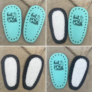 May include: Four pairs of baby shoe soles, two pairs are turquoise blue with black stitching and the text "but first milk" with a baby bottle graphic, and two pairs are black with white fleece lining.