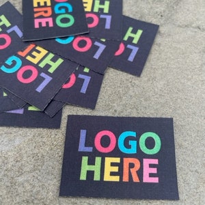 May include: A collection of rectangular black stickers featuring the text "LOGO HERE" printed in a vibrant, multi-colored font. Each letter of the text displays a different color, including blue, pink, green, orange, and purple, arranged in a stacked layout.