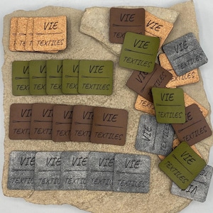 May include: A collection of square labels in tan, olive green, brown, and grey, each featuring the text VIE TEXTILES and a needle graphic. The labels are arranged in organised rows and scattered piles, showcasing varied textures and colours.