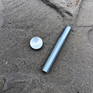 May include: A small, silver metal tool with a textured handle and a circular, flat head. The tool is lying on a rough, grey stone surface. The tool appears to be a punch or a similar type of hand tool.