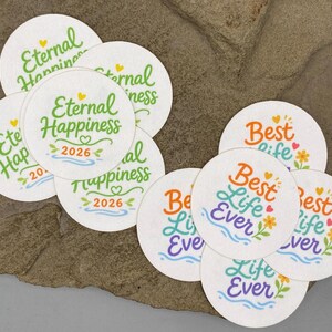 May include: A collection of circular white stickers featuring two distinct designs. One design displays the green text Eternal Happiness with the year 2026, while the other features the colorful text Best Life Ever with floral and heart accents.