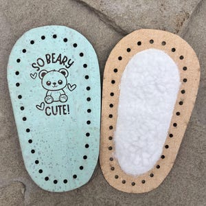 Könnte beinhalten: Zwei ovale Schuhsohlen mit vorgestanzten Löchern entlang der Ränder. Die linke Sohle ist hellminzgrün mit gesprenkelter Textur und einer schwarzen Bärengrafik mit dem Text SO BEARY CUTE. Die rechte Sohle ist hellbraun mit einem weißen Kunstfellfutter.