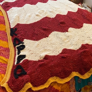 Op de afbeelding: Een gehaakt deken met een chevron-patroon in wit en bordeauxrood met een gouden rand. Het deken heeft het woord "Kappa" in zwart garen aan de zijkant.