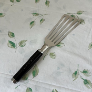 Puede incluir: Una espátula ranurada de acero inoxidable con mango negro. La espátula tiene una cabeza rectangular con cinco ranuras. El mango es negro con un acento plateado en la base. La espátula está sobre un fondo blanco con patrones de hojas verdes.