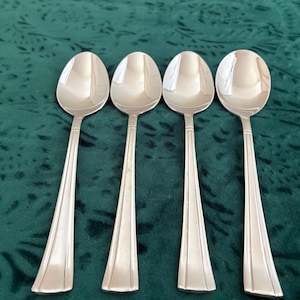 May include: A set of four identical silver-toned metal spoons arranged in a row. Each spoon features a smooth, oval-shaped bowl and a long, slender handle with three vertical embossed lines running down the center, ending in a flared base.