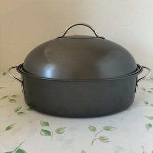 May include: A dark gray oval roasting pan with a matching lid and silver handles. The pan is sitting on a white tablecloth with a green leaf pattern. The lid has a silver handle.