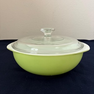May include: A round, lime green casserole dish with a clear glass lid and a small, clear glass handle. The dish has white handles on either side. The lid sits on a white rim. The dish is likely made of glass.