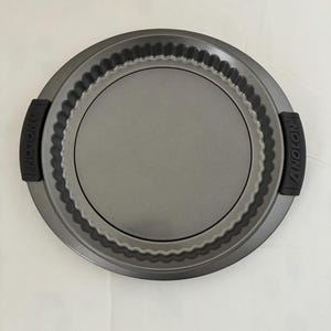 Puede incluir: Un molde para hornear redondo y gris con borde estriado y asas negras. El molde tiene una superficie antiadherente y está diseñado para hornear tartas. El nombre de la marca "ANOLON" está impreso en las asas. Diámetro: aprox. 25 cm.