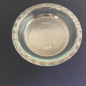 Op de afbeelding: Een heldere glazen taartvorm met een gegolfde rand. De vorm heeft een diameter van 9,5 inch en heeft de tekst "100 Years 1915" en "Pyrex PL USA" gegraveerd op de bodem.