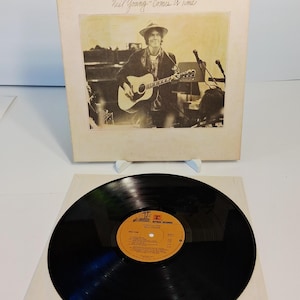 Puede incluir: Un álbum de vinilo de Neil Young, titulado "Comes A Time". La portada del álbum presenta una imagen en tonos sepia de Neil Young tocando una guitarra acústica. El disco en sí es negro con una etiqueta amarilla y naranja.