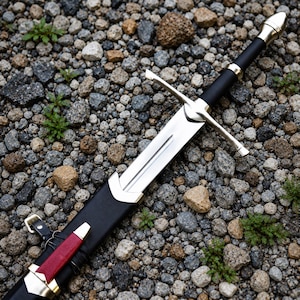 May include: A silver-coloured metal sword with a black handle and a matching black scabbard. The scabbard features a silver-coloured metal tip and a red leather accent piece with a small silver buckle strap attached to the side of the sheath.